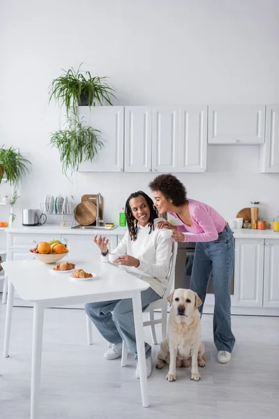 Sorrindo afro-americano mulher abraçando namorado com smartphone perto do café da manhã e labrador na cozinha — Fotografia de Stock