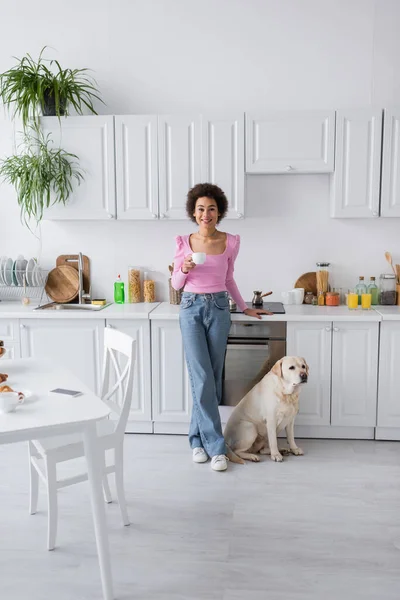 Mujer afroamericana sonriente sosteniendo taza de café cerca de labrador en la cocina - foto de stock