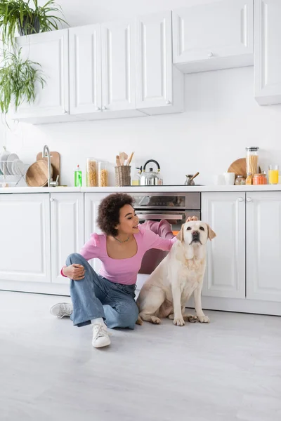 Sorridente donna afro-americana che accarezza labrador sul pavimento in cucina — Foto stock