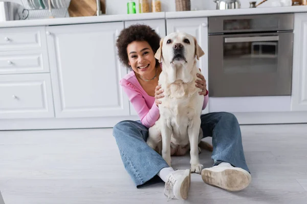 Young african american woman petting labrador on floor in kitchen — Fotografia de Stock