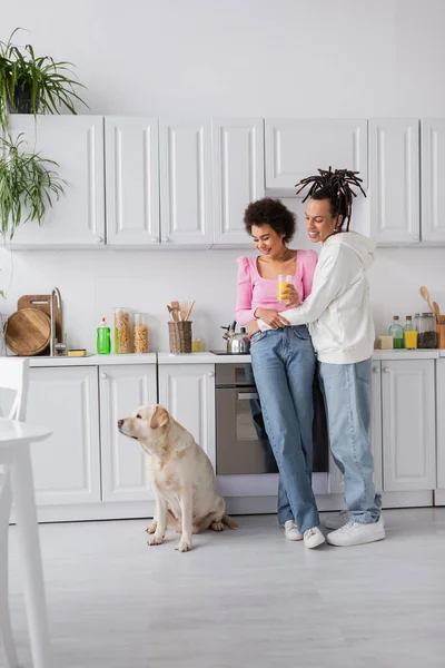 Souriant couple afro-américain avec du jus d'orange regardant labrador dans la cuisine — Photo de stock