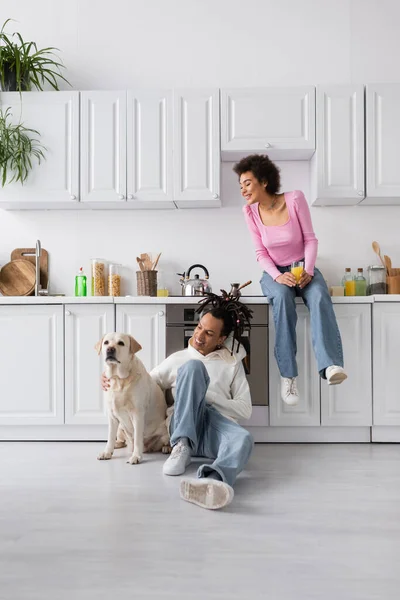 Sourire femme afro-américaine tenant du jus d'orange près de petit ami avec labrador à la maison — Photo de stock