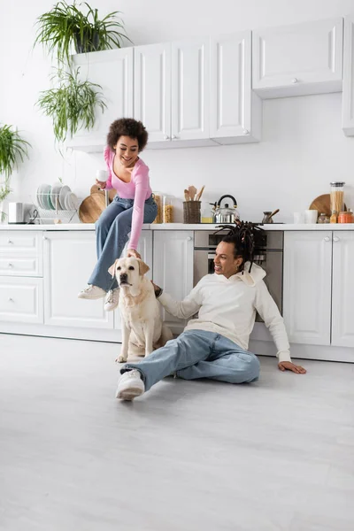 Cheerful african american woman holding coffee near boyfriend and labrador on floor in kitchen - foto de stock