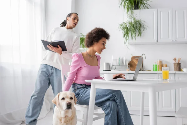 African american woman using laptop near boyfriend with notebook and labrador in kitchen — Stock Photo