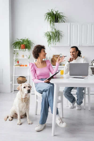 Smiling african american woman with notebook talking to boyfriend near devices and labrador at home — Stock Photo