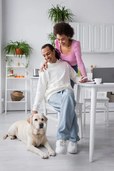 Smiling african american woman hugging boyfriend near labrador and devices in kitchen — Stock Photo