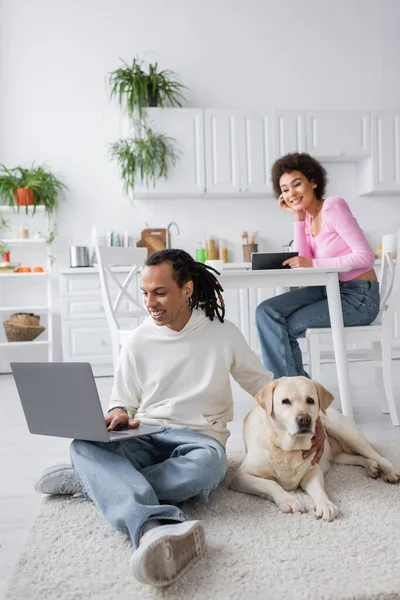 Sonriente pareja afroamericana usando portátil cerca de labrador acostado en la alfombra en casa - foto de stock