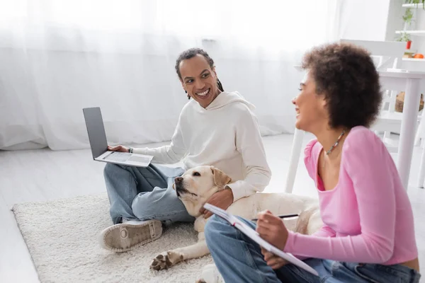 Cheerful african american couple with laptop talking near labrador on carpet — Photo de stock