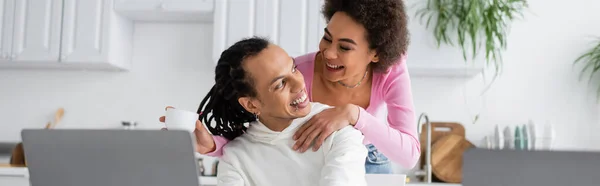 Smiling african american woman holding coffee and hugging boyfriend in kitchen, banner — Photo de stock