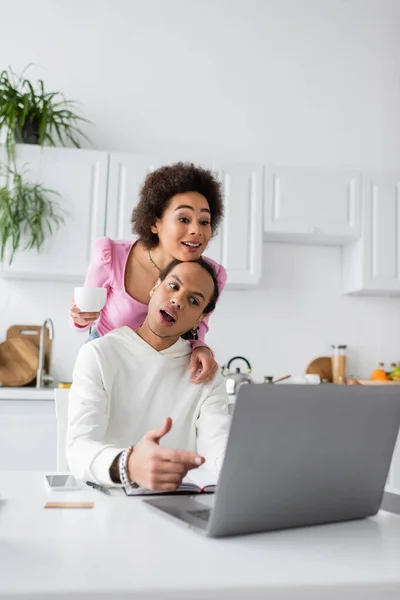 African american woman holding coffee while boyfriend pointing at laptop at home — Fotografia de Stock