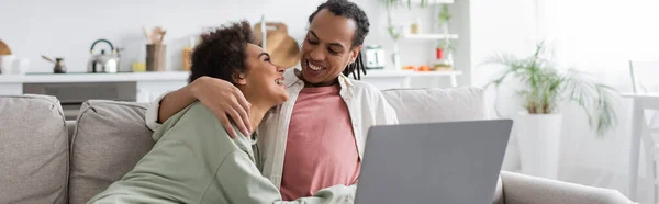 Sorridente donna afro-americana che abbraccia il fidanzato vicino al computer portatile sul divano a casa,, banner — Foto stock