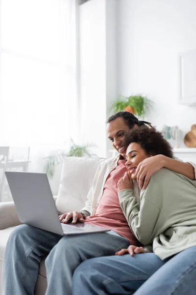 Sorrindo casal afro-americano abraçando ao usar laptop em casa — Fotografia de Stock