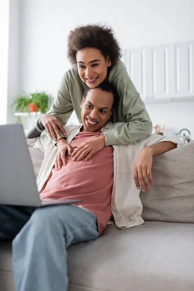 Afro americano donna abbracciare tatuato fidanzato vicino computer portatile su divano a casa — Foto stock