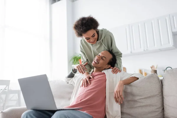 Smiling african american woman hugging tattooed boyfriend with laptop on couch at home — Photo de stock