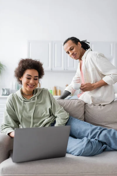 Femme afro-américaine gaie utilisant un ordinateur portable près du petit ami avec du café à la maison — Photo de stock