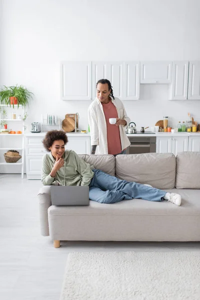Giovane donna afroamericana che guarda il computer portatile vicino al fidanzato con una tazza di caffè a casa — Foto stock