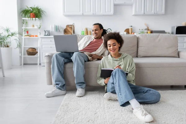 Sourire femme afro-américaine écrit sur un ordinateur portable près de petit ami avec ordinateur portable à la maison — Photo de stock