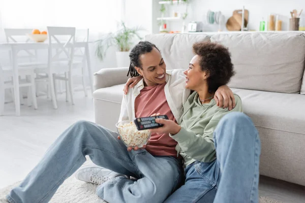 Positive african american couple with popcorn hugging while watching movie at home — Stock Photo