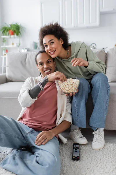 African american woman holding popcorn near boyfriend and remote controller at home — Stock Photo