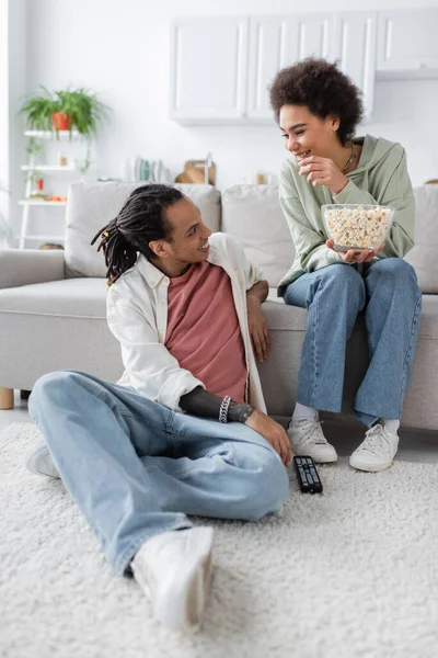 Positiva pareja afroamericana con palomitas de maíz y controlador remoto hablando en casa - foto de stock