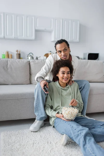 Sonriente pareja afroamericana viendo películas y sosteniendo palomitas de maíz en casa - foto de stock