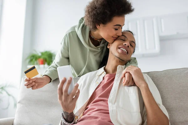 Smiling african american woman holding credit card and kissing boyfriend with smartphone at home — Stock Photo