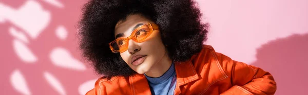 Mujer americana africana rizada en gafas de sol naranjas elegantes mirando hacia otro lado en rosa, bandera - foto de stock