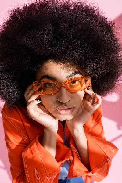 Vista aerea di ricci donna africana americana in eleganti occhiali da sole arancioni guardando la fotocamera su rosa — Foto stock