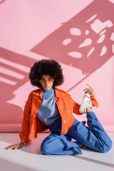 Longitud completa de la joven mujer afroamericana en gafas de sol naranja con estilo y traje de moda posando en rosa - foto de stock