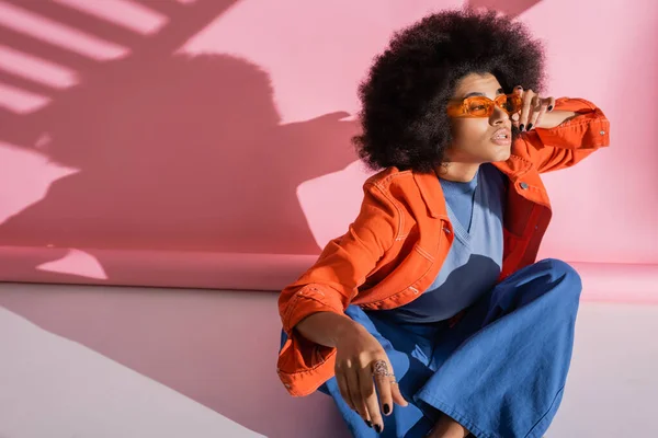Curly african american woman in trendy outfit adjusting orange sunglasses on pink — Stockfoto