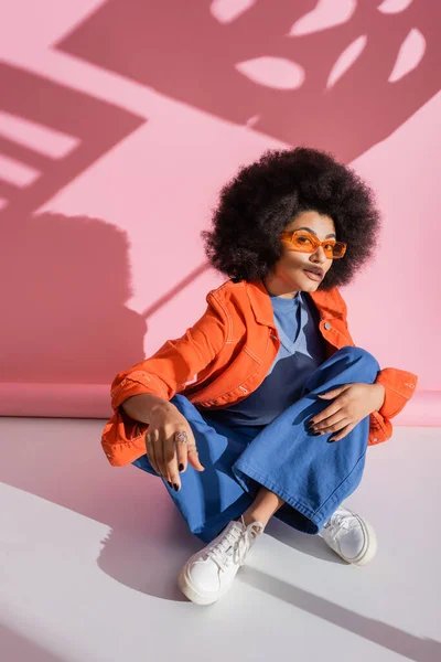 Curly african american woman in stylish orange sunglasses and trendy outfit sitting on pink — Stockfoto