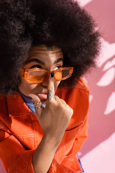 Vue grand angle de femme afro-américaine bouclée ajustant les lunettes de soleil orange tendance sur rose — Photo de stock