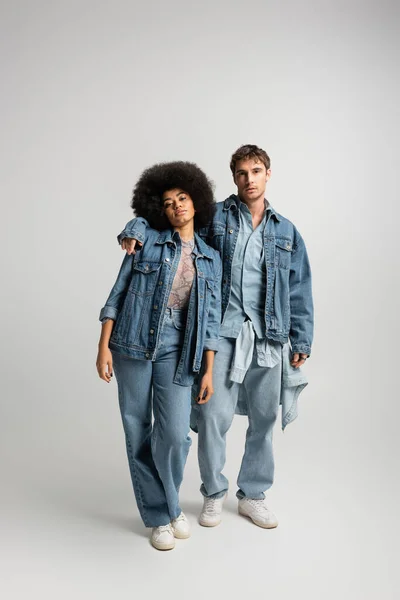 Full length of young man in denim outfit hugging african american woman on grey — Stock Photo
