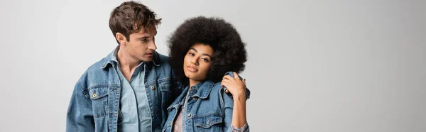 Young man in denim outfit hugging curly african american woman isolated on grey, banner — Stock Photo