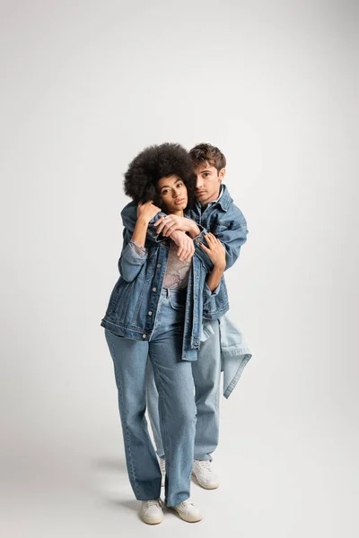 Full length of young man in denim outfit hugging curly african american woman on grey — Stock Photo
