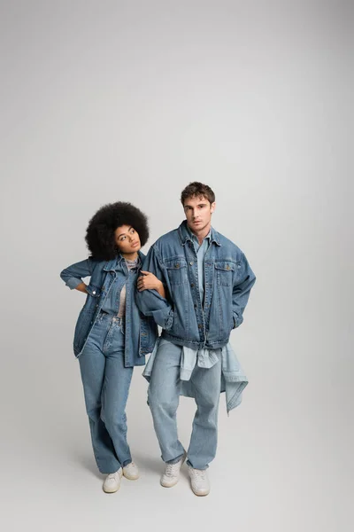 Full length of young man in denim outfit posing with hands in pockets near curly african american woman on grey — Fotografia de Stock