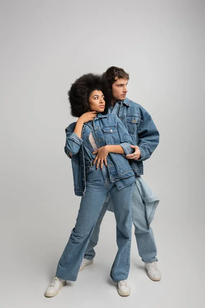 Full length of young multiethnic couple in denim outfit hugging and looking away on grey — Stock Photo