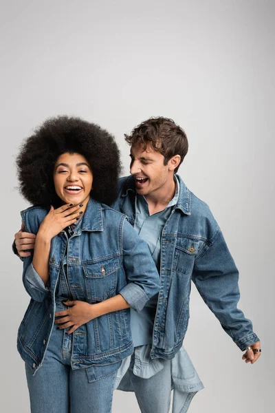 Glückliche multiethnische Mann und Frau in Jeans-Outfit lachen isoliert auf grau — Stockfoto