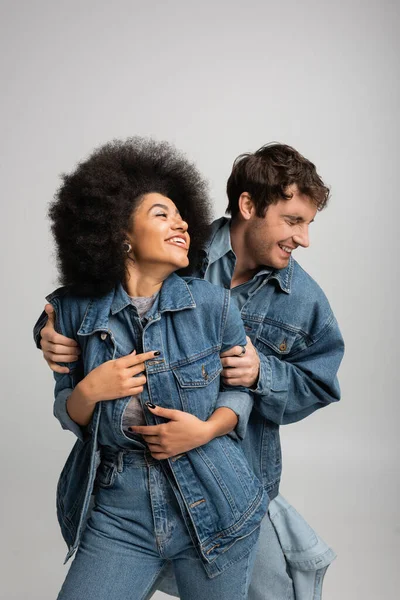 Веселий багатоетнічний чоловік і жінка в джинсовому вбранні сміється ізольовано на сірому — стокове фото