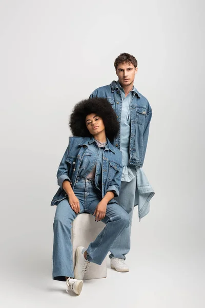 Full length of curly african american woman sitting near man in denim outfit on grey — Fotografia de Stock