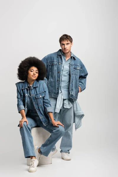 Full length of curly african american woman sitting near man in denim outfit posing with hands in pockets on grey — Stock Photo