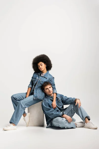 Full length of brunette african american woman sitting with man in denim outfit on grey — Stock Photo