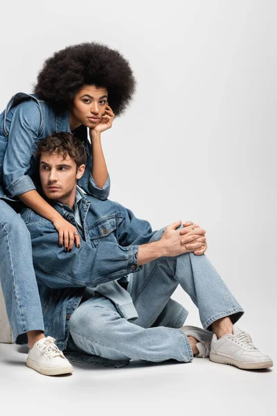 Brunette african american woman in sneakers sitting with man in denim outfit on grey - foto de stock