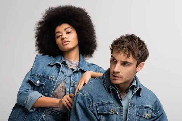 Brunette african american woman looking at camera and leaning on man in denim outfit isolated on grey — Stock Photo