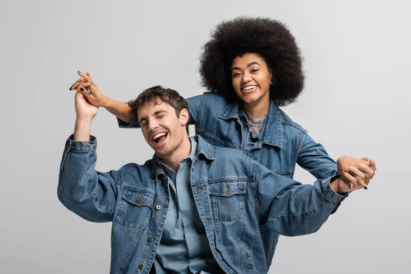 Cheerful african american model looking at camera and holding hands with stylish man isolated on grey — Stock Photo