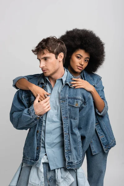 Curly african american model looking at camera while embracing stylish man in denim clothes isolated on grey — Stock Photo