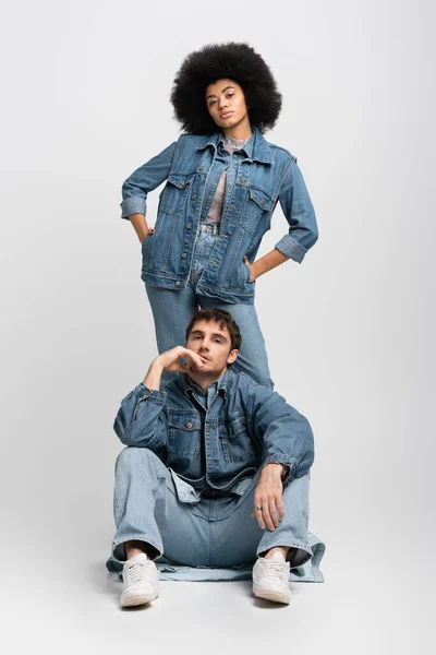 Full length of curly african american woman standing with hands in pockets behind man in denim outfit on grey — Stock Photo