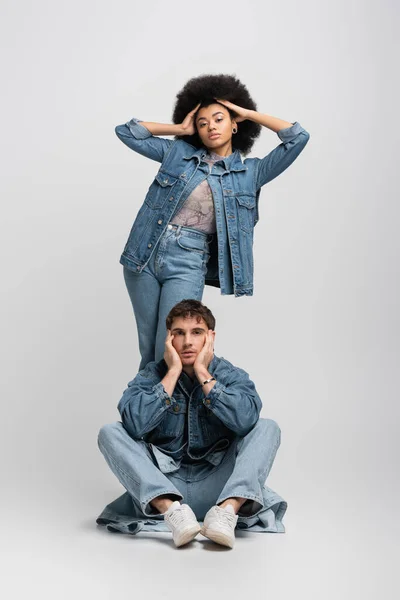 Full length of curly african american woman posing behind man in denim outfit and sneakers sitting on grey — Stock Photo