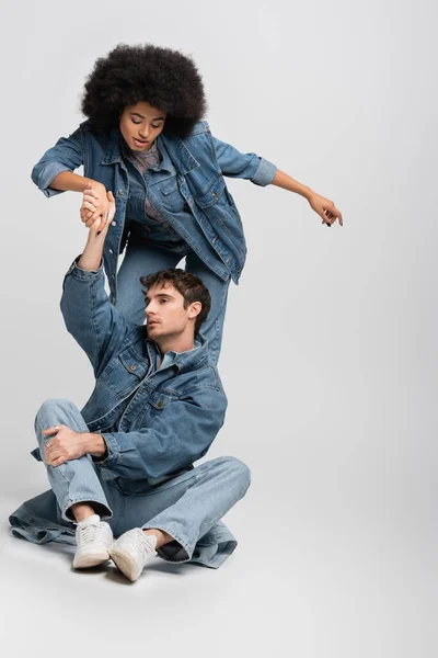Full length of man in denim clothes sitting and holding hand of curly african american woman on grey — Stock Photo