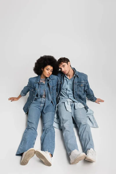 Full length of stylish interracial couple in denim clothes sitting in sneakers on grey — Stock Photo